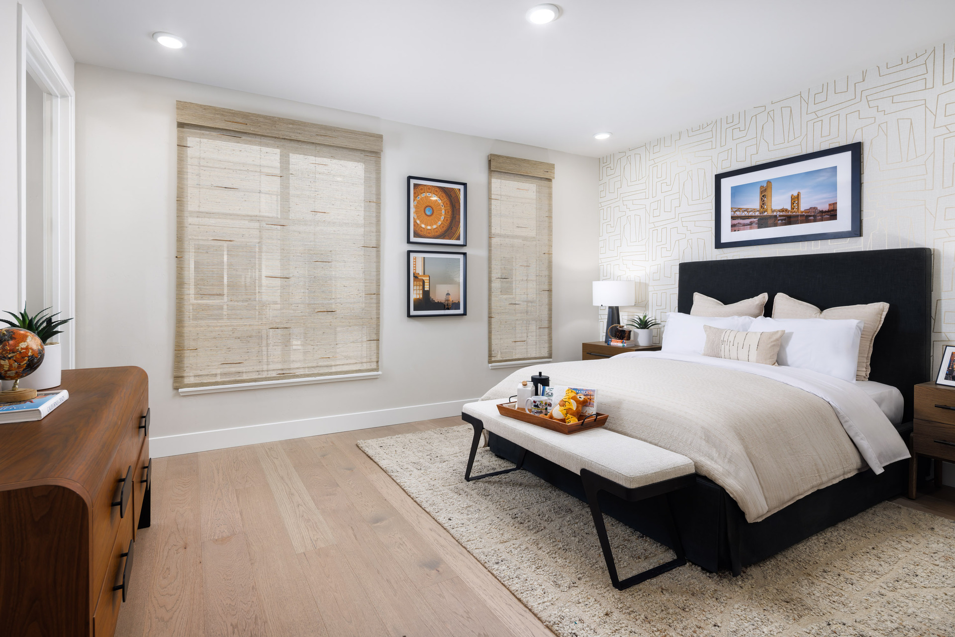 bedroom with light wood floors, two windows, black upholstered bed with white & tan bedding, and wood dresser