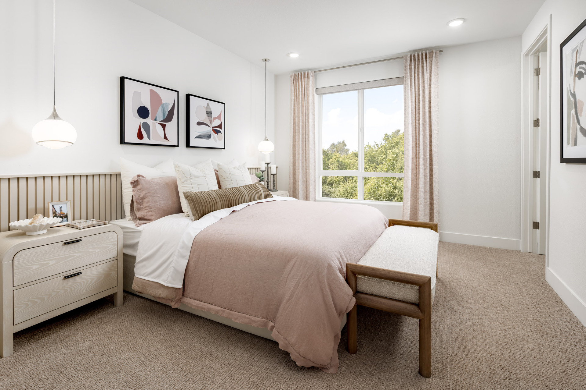bedroom with tan carpet, blush pink & white bedding, wood bench at foot of bed, and window