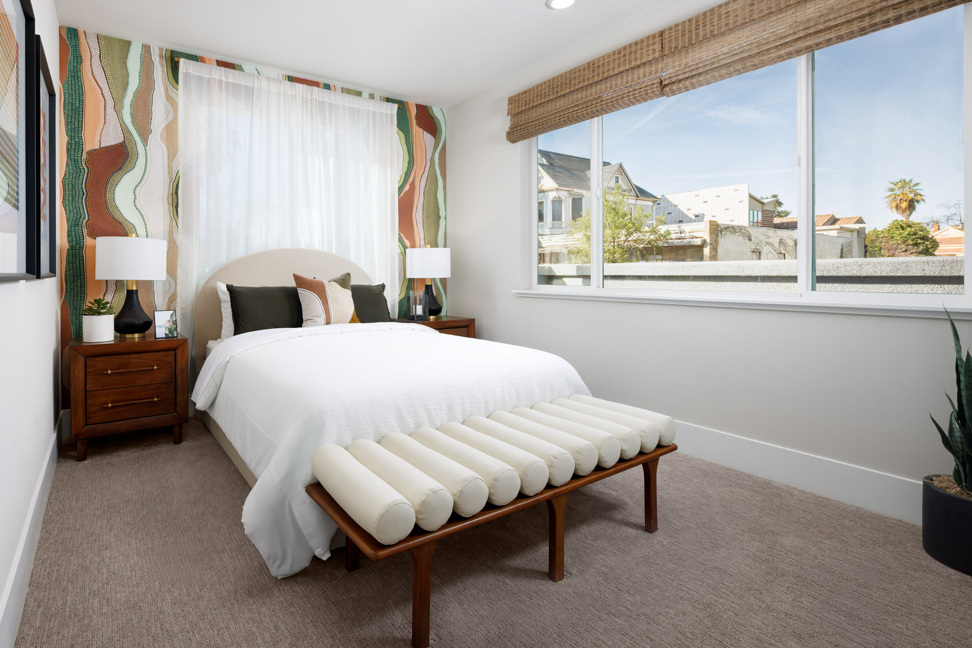 bedroom with tan carpet, colorful wallpaper behind bed, wood nightstands, and large window with views of downtown