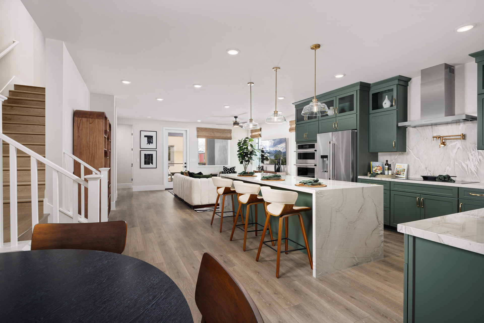 kitchen with wood floors, green stacked cabinets, waterfall quartz counter at island, stainless steel hood above stove, and view of great room