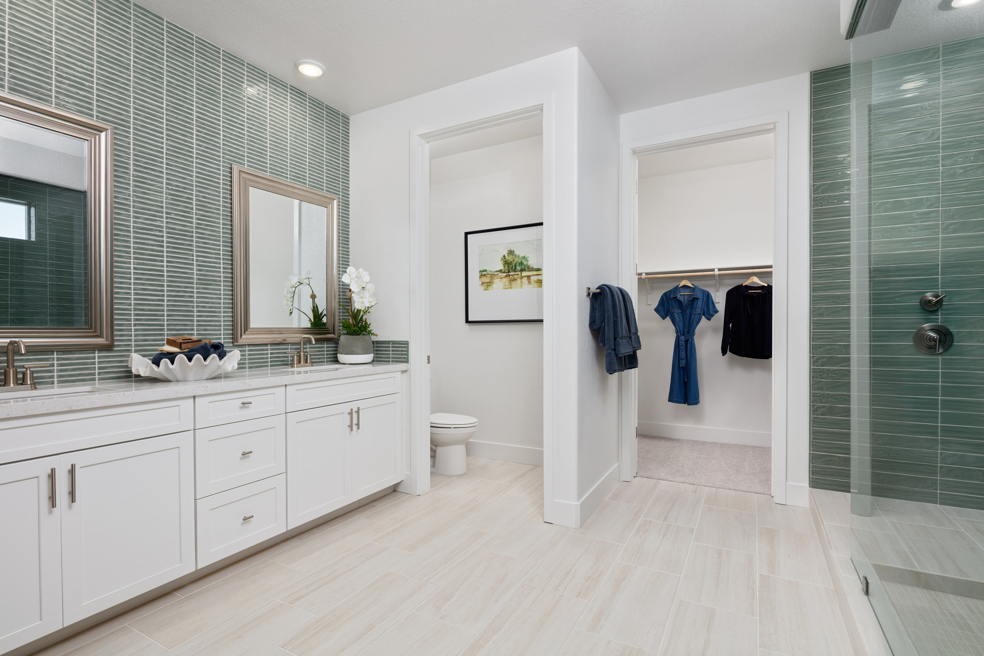 bathroom with green tile on wall at vanity & shower, light tan tile floors, white cabinets, two sinks, and walk-in closet