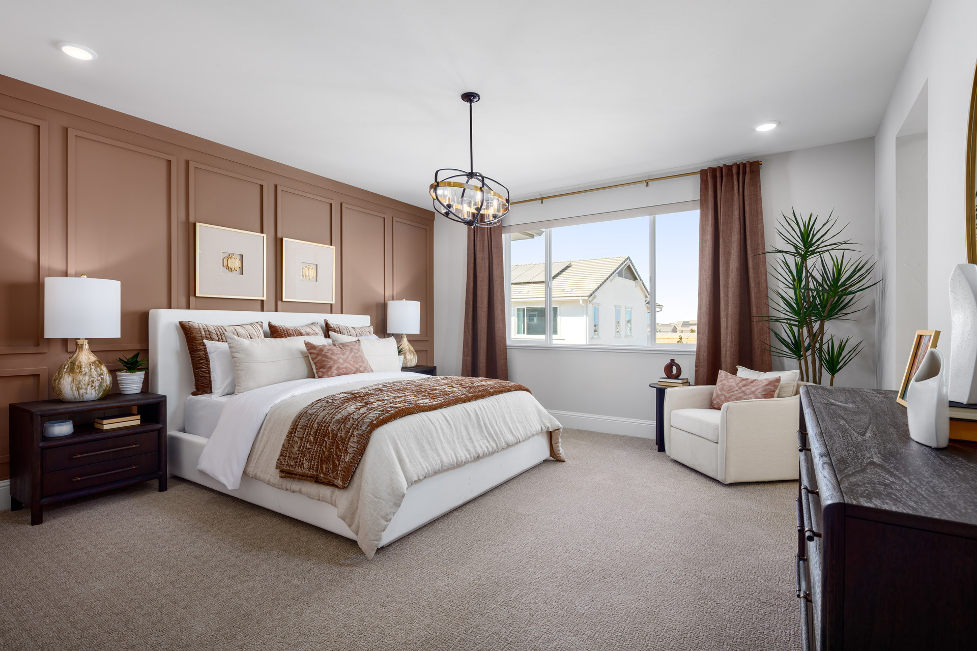 bedroom with brown wall behind white upholstered bed, dark wood nightstands & dresser, tan carpet, large window with brown panels, and white accent chair in corner