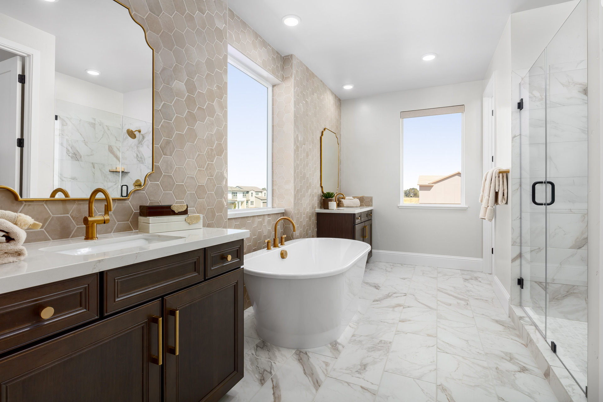 bathroom with tan hexagon wall tile, espresso cabinets, two vanities, walk-in shower, and window above large freestanding soaking tub